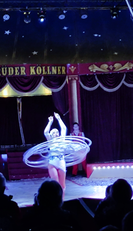 Eine Künstlerin performt mit Hula-Hoops in einem Zirkus. Zuschauer sind im Vordergrund.